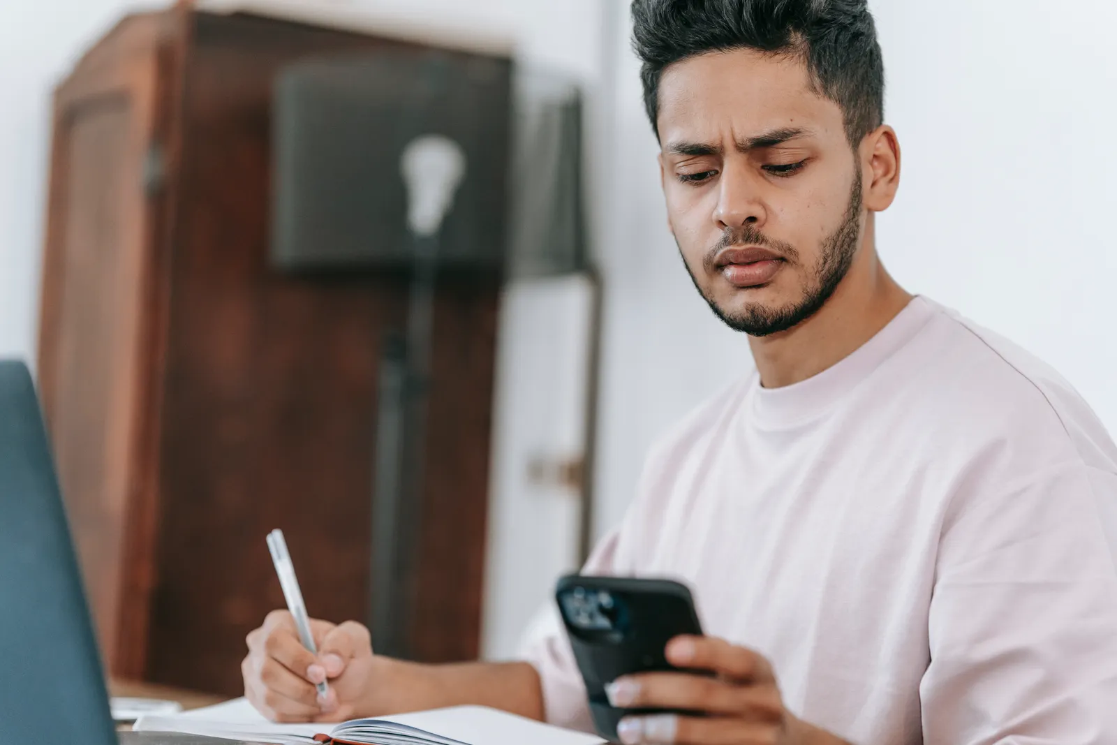 Homem consultando o celular e fazendo anotações ao lado de um notebook
